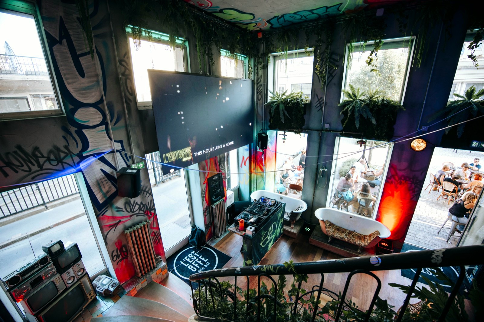 La salle vue depuis la mezzanine : murs graffés, plantes suspendues, baignoires détournées en banquettes et platines au centre